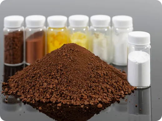 A pile of brown mineral granules in front of sample vials holding powders in brown, orange and white