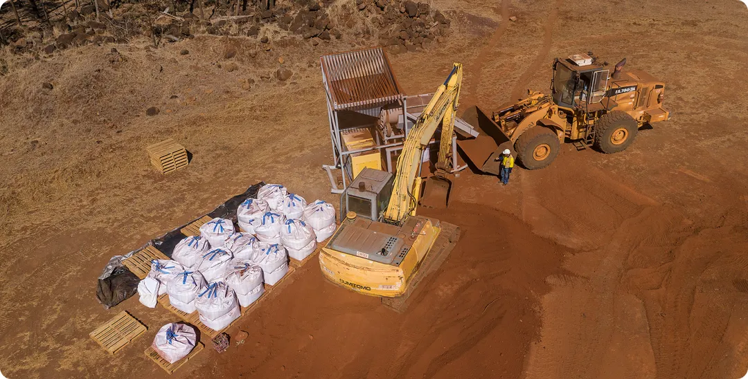 Aerial view of an excavator and front-end loader working red-earth ground beside pallets of bulk sample bags
